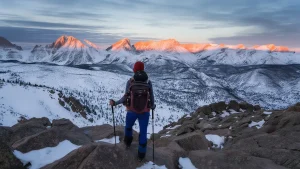 A hiker stands on a snowy ridge, overlooking sunlit mountain peaks.